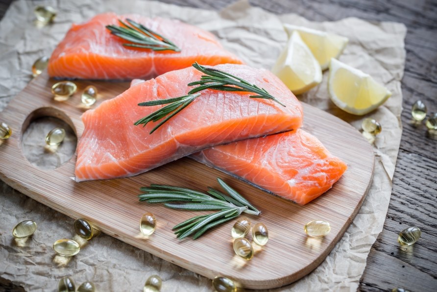 Three salmon fillets on a cutting board with omega-3 capsules and lemon wedges.